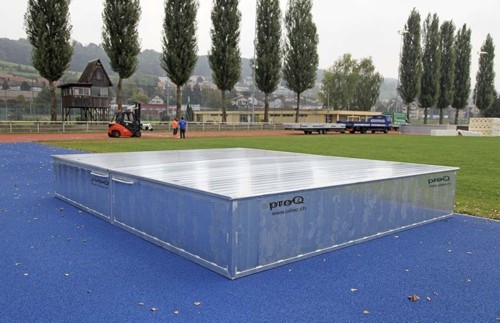 Ein großer, rechteckiger Schutzcontainer von proQ steht auf einem blauen Sportplatz, umgeben von Bäumen und Gebäuden.