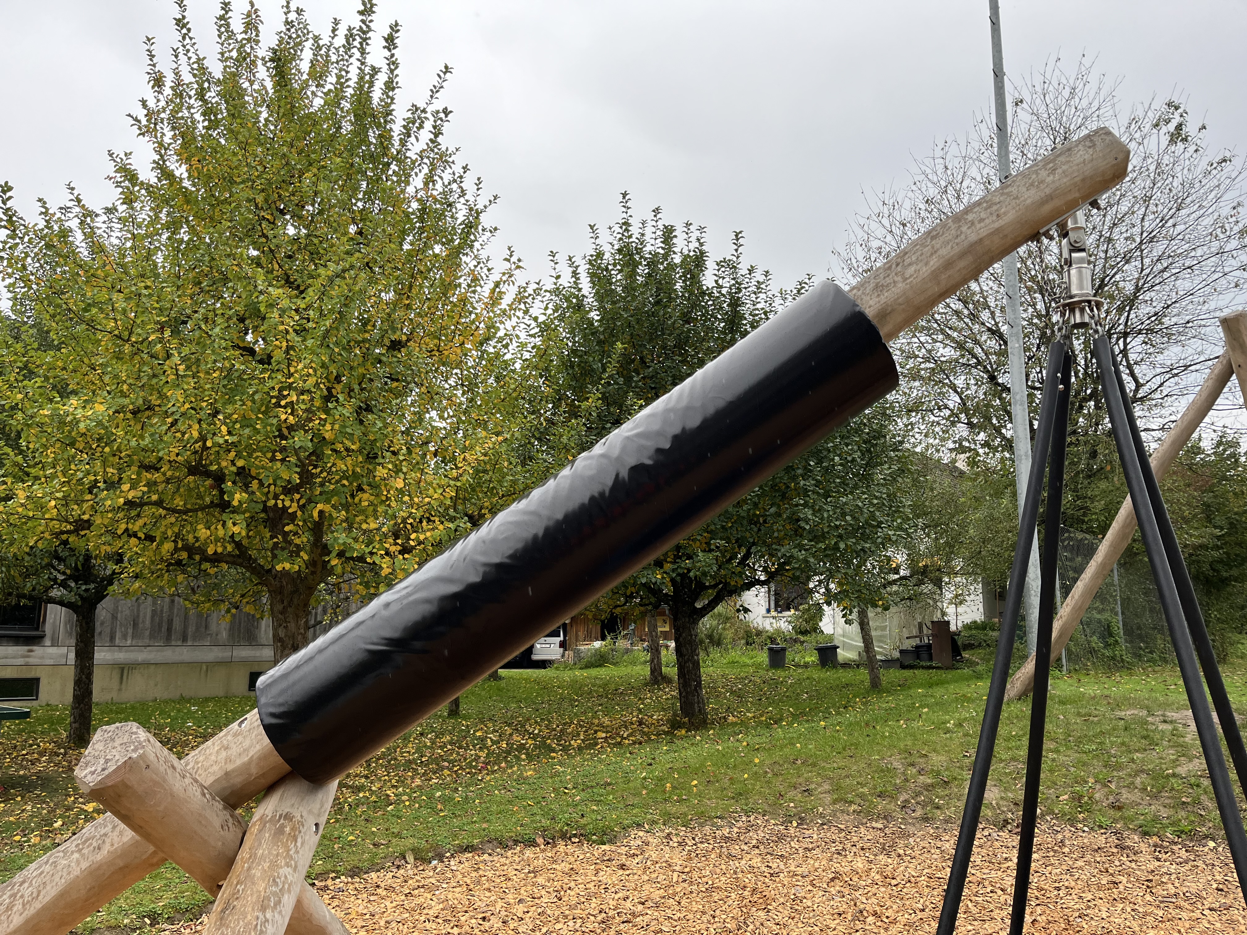 Nahaufnahme eines Holzspielzeugs mit schwarzem Kunststoffüberzug im Freien, umgeben von herbstlichen Bäumen und Holzchips am Boden.