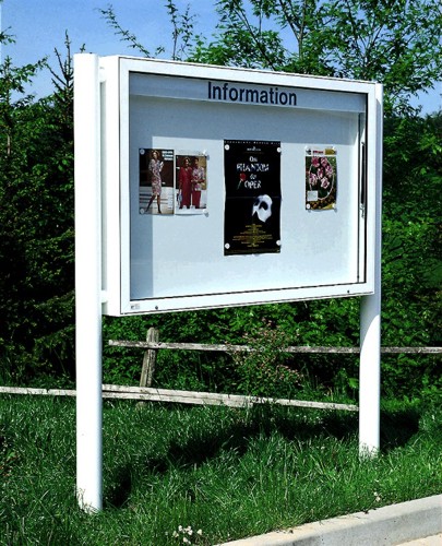 Ein weißes Informationsschild im Freien, darin befinden sich mehrere Plakate. Es ist von grünem Gras und Bäumen umgeben.