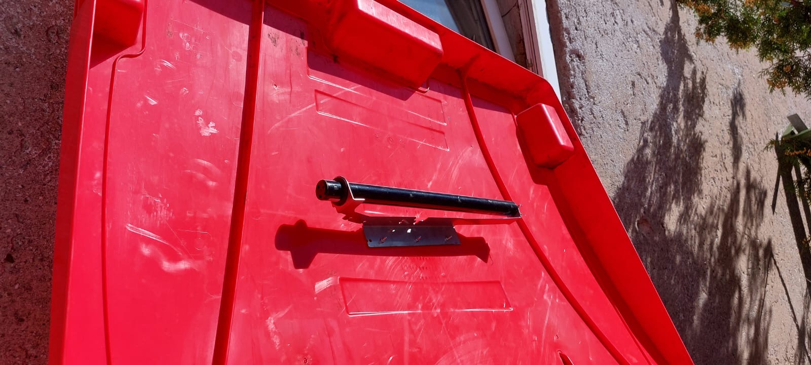 Detailaufnahme einer roten Containerabdeckung mit einem schwarzen Griff und Metallbefestigungen, die an einer Außenwand lehnt.