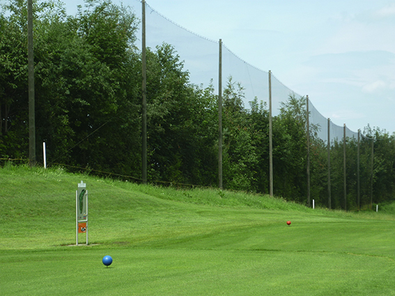 Ansicht eines Golfplatzes mit blauen und roten Abschlagsmarkierungen sowie einem hohen Abgrenzungsnetz entlang des Fairways.