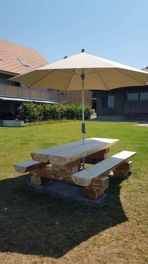 Ein Holz-Picknicktisch mit zwei Bänken und einem großen Sonnenschirm auf einer grünen Rasenfläche vor einem Gebäude mit Holzfassade.