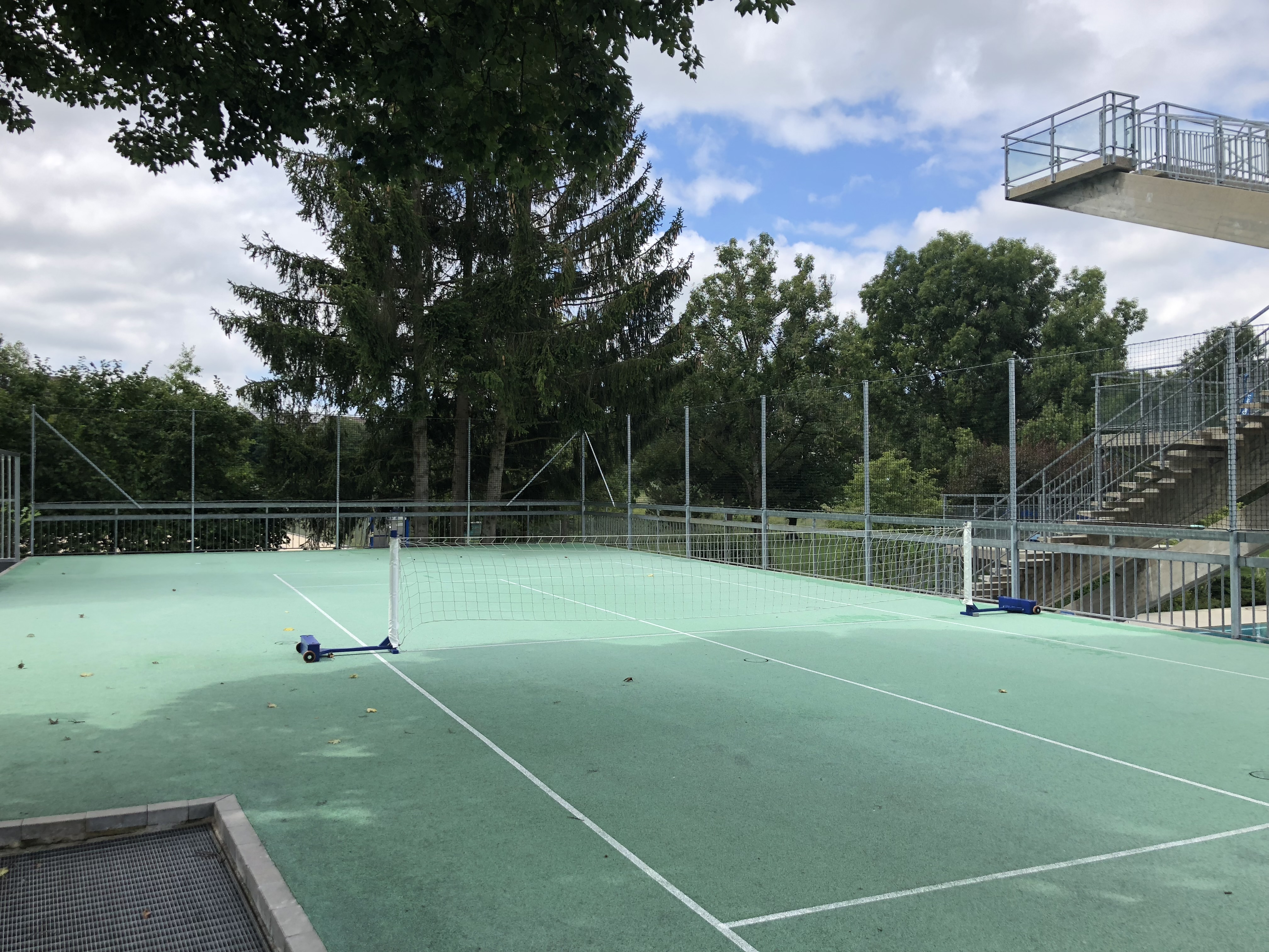 Ein leerer Tennisplatz im Freien, umgeben von hohen Bäumen und einem Sprungturm im Hintergrund bei bewölktem Himmel.