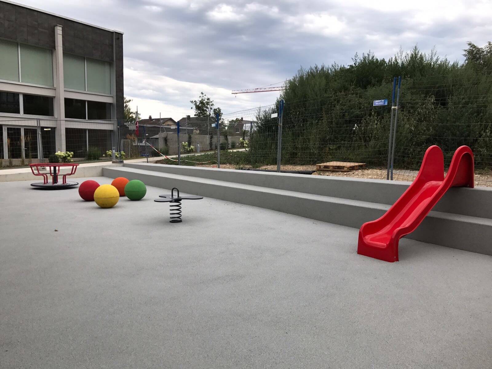 Une aire de jeux moderne avec un toboggan rouge, des balles colorées, un carrousel et une bascule sur un revêtement de sol sûr. Idéal pour que les enfants puissent jouer en plein air.
