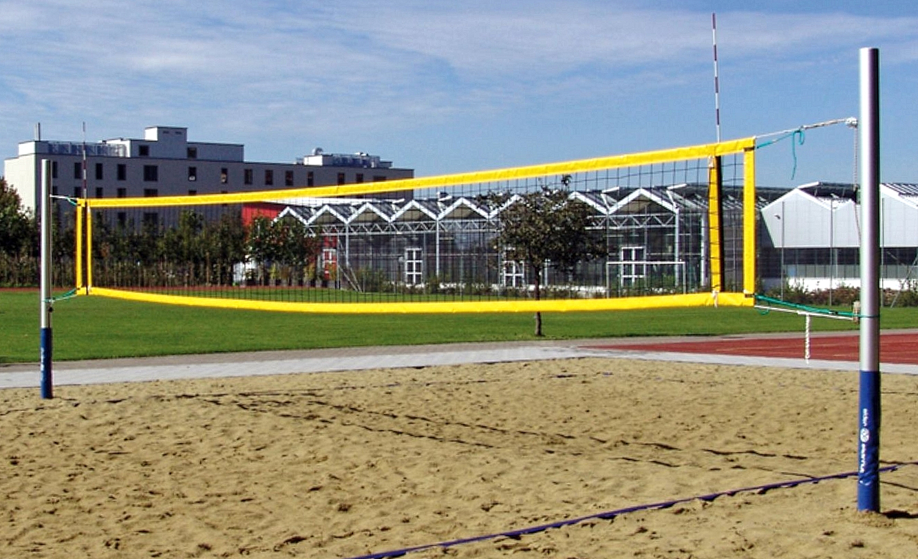 Ein Strand-Volleyballplatz mit gelbem Netz auf sandigem Boden, umgeben von Grünflächen und Gebäuden im Hintergrund.