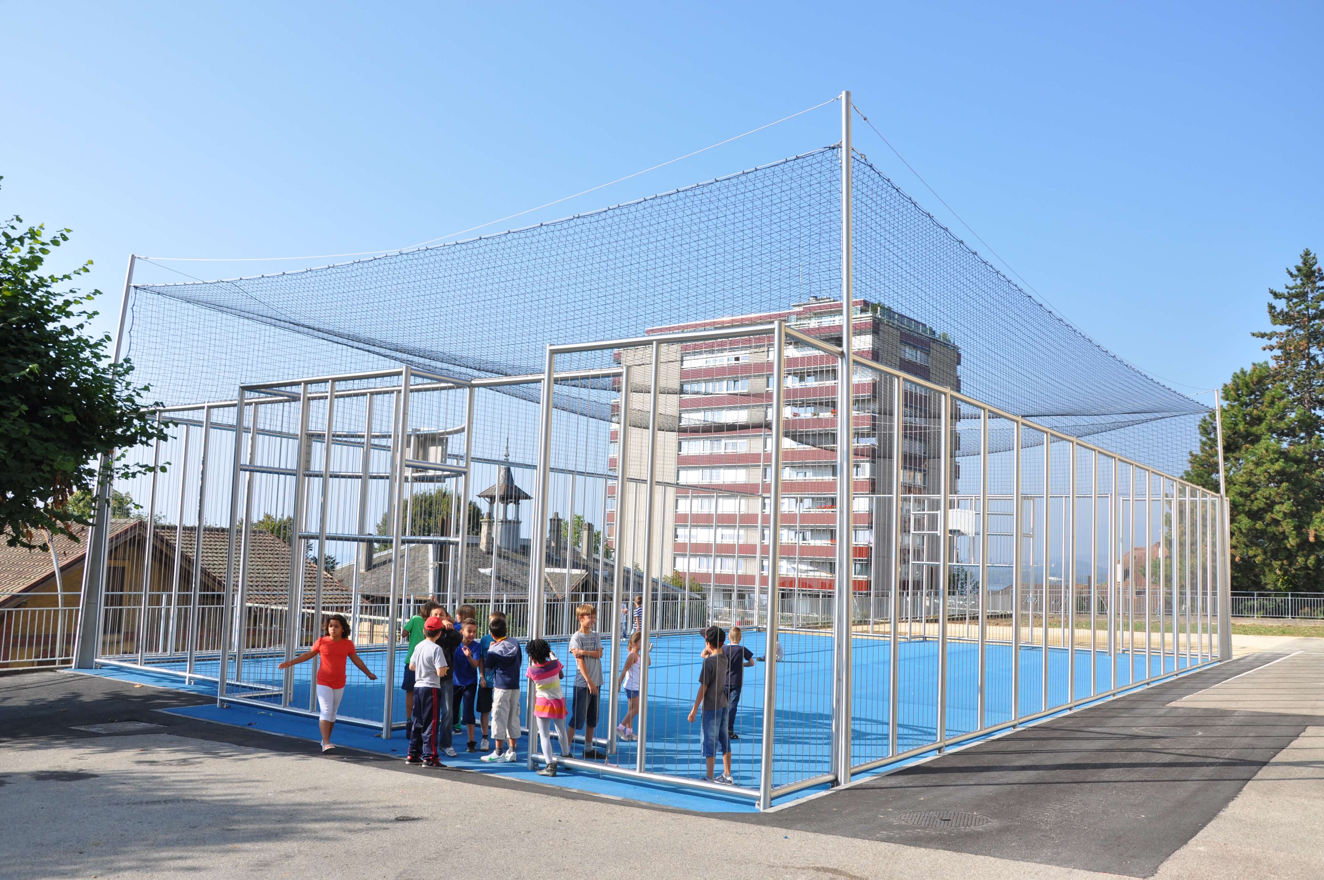 Un groupe d'enfants joue en plein air sur un terrain de sport bleu clôturé, entouré de bâtiments