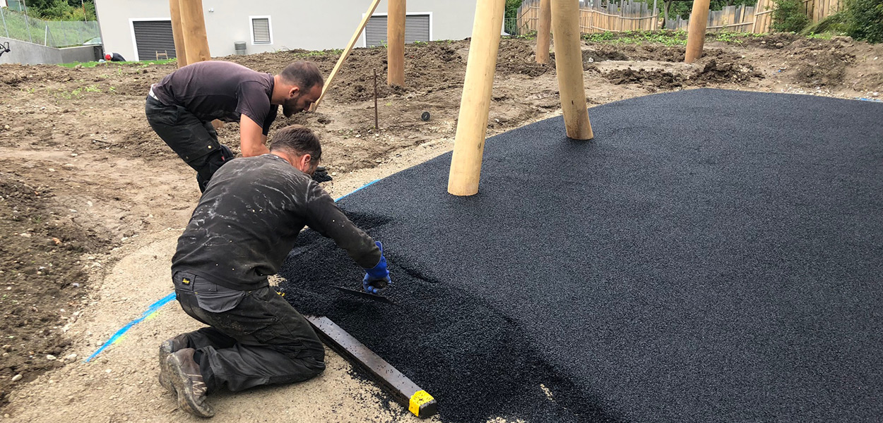 Zwei Männer installieren einen schwarzen Gummibelag auf einem Spielplatz, umgeben von unbebautem Gelände und Holzstrukturen