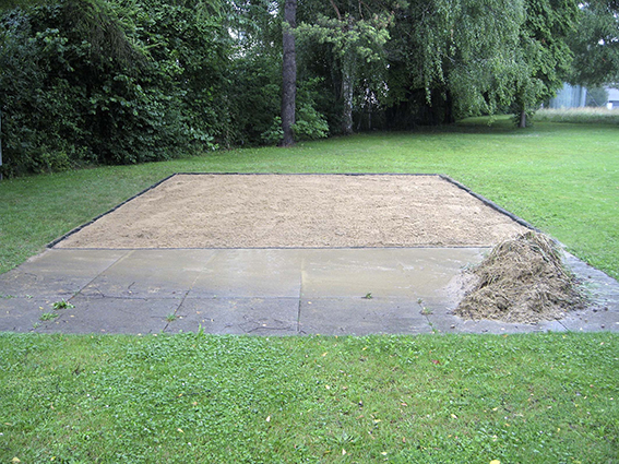 Une zone rectangulaire dans le jardin a été préparée avec du sable, entourée d'herbe et d'arbres.