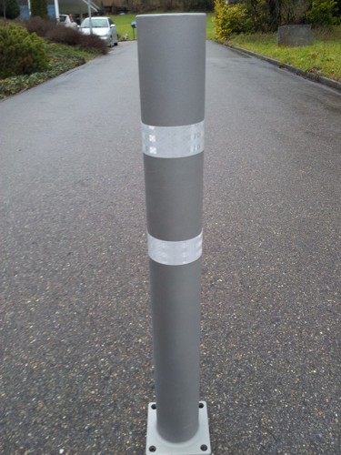 Ein grauer Verkehrspoller steht auf einem asphaltierten Boden, umgeben von Grünflächen.