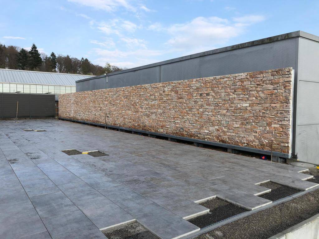 Un espace extérieur moderne avec une façade en pierre et des dalles de béton grises, entouré par la nature et des bâtiments en arrière-plan.