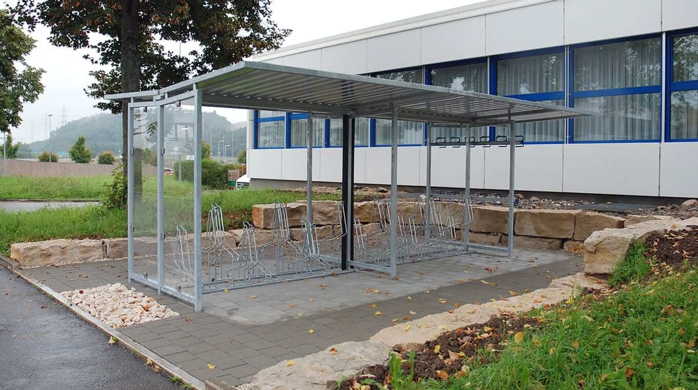 Un abri à vélos moderne en métal avec un toit, situé à côté d'un immeuble de bureaux, entouré d'espaces verts.