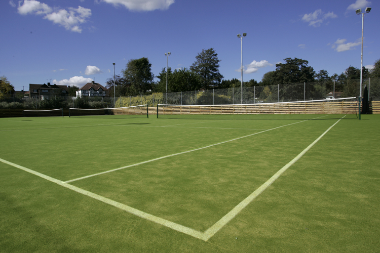 Un court de tennis vide avec du gazon synthétique vert sous un ciel bleu et quelques nuages, entouré d'arbres et de clôtures.