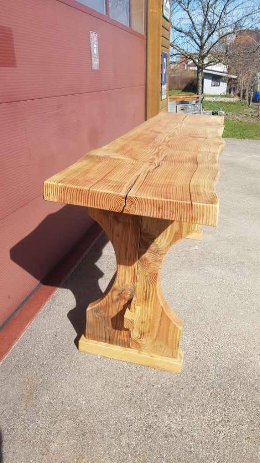 Ein massiver Holztisch mit natürlicher Holzmaserung steht im Freien an einer Hauswand.