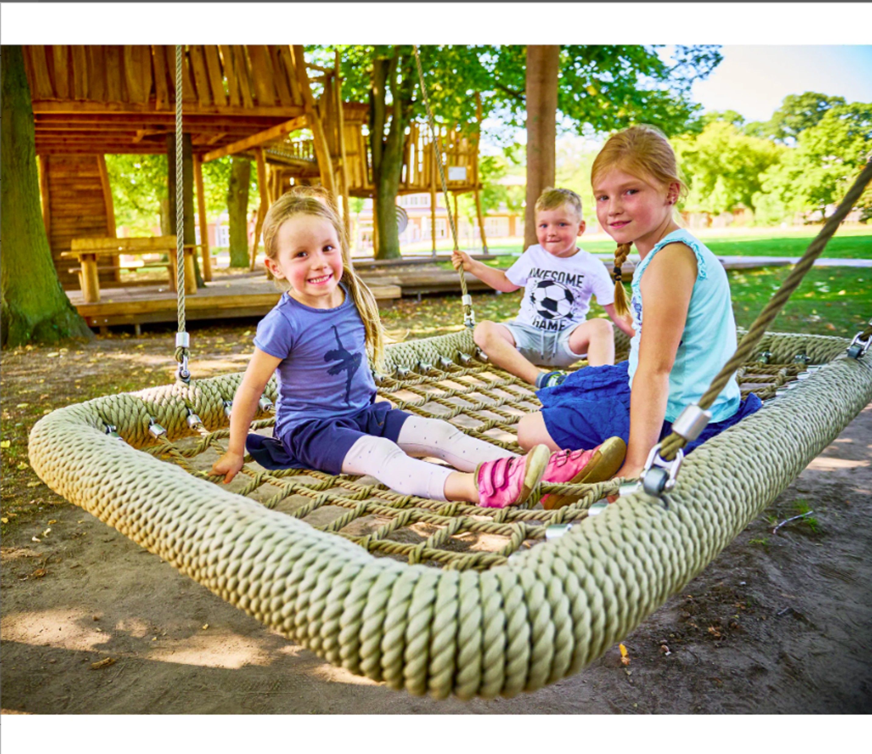 Trois enfants sont assis en riant sur une grande balançoire rectangulaire en corde sur une aire de jeux avec un portique en bois en arrière-plan