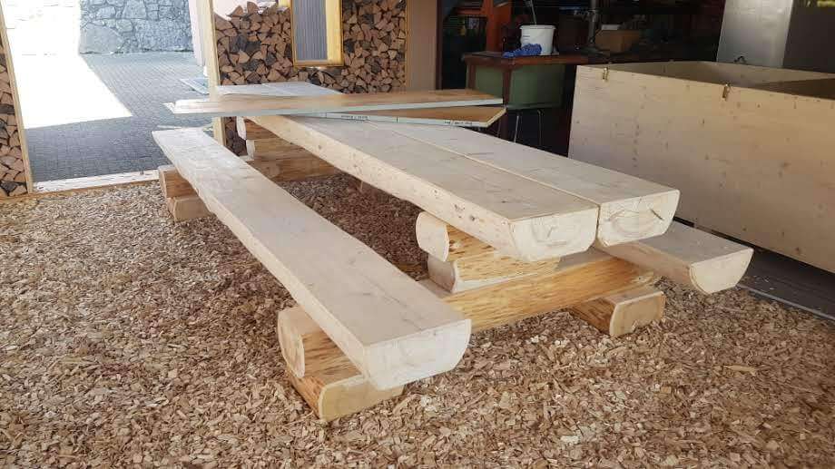 Große, robuste Holzbänke und ein Tisch aus dicken Balken auf einer Schicht Holzspäne in einer Werkstattumgebung