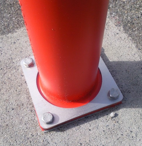 Eine rote Metallstange mit einer rechteckigen Befestigungsplatte, die mit vier Bolzen auf einem Betonboden fixiert ist.