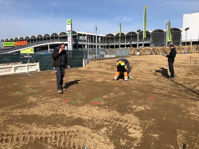 Trois personnes au travail sur un chantier devant un centre commercial sous un ciel bleu éclatant