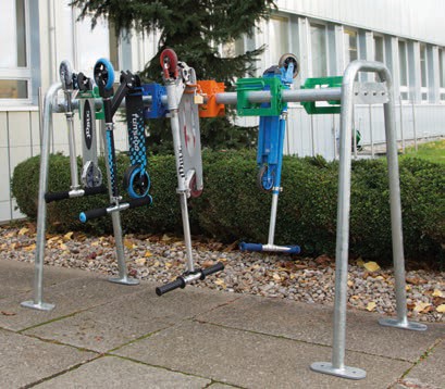 Fünf bunte Scooter sind an einem stabilen Fahrradständer im Freien nebeneinander geparkt, mit Hintergrund aus Hecken und einem Gebäude.