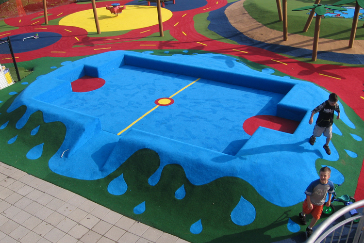 Deux enfants jouent sur une aire de jeux colorée avec un élément d'escalade bleu en forme de terrain de jeu, entouré de chemins colorés et d'un sol vert.
