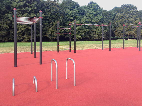 Ein moderner Outdoor-Fitnessbereich mit Klettergerüsten und Stangen auf rotem Gummiboden, umgeben von Rasen und Bäumen.
