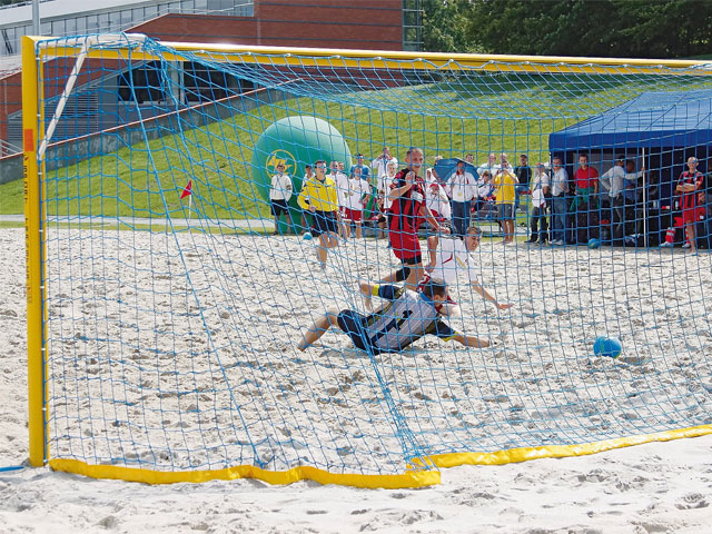 Spieler schießt Tor während eines Beachsoccer-Spiels auf einem sandigen Spielfeld, umgeben von Zuschauern und einem Torwart im Netz.