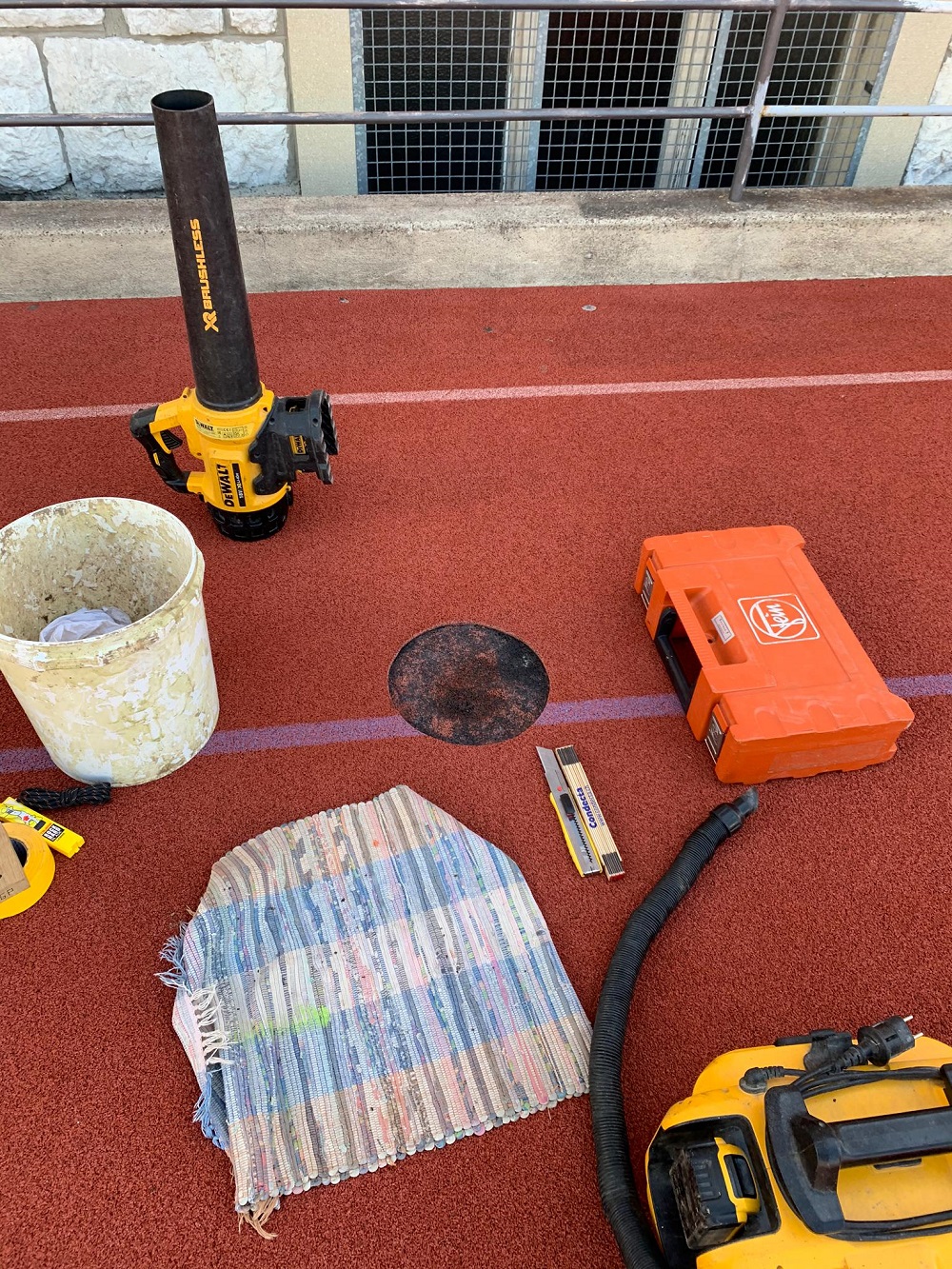 Sur une piste rouge se trouvent différents outils et matériaux tels qu'un souffleur de feuilles, un aspirateur, un tapis, un niveau à bulle et un seau.