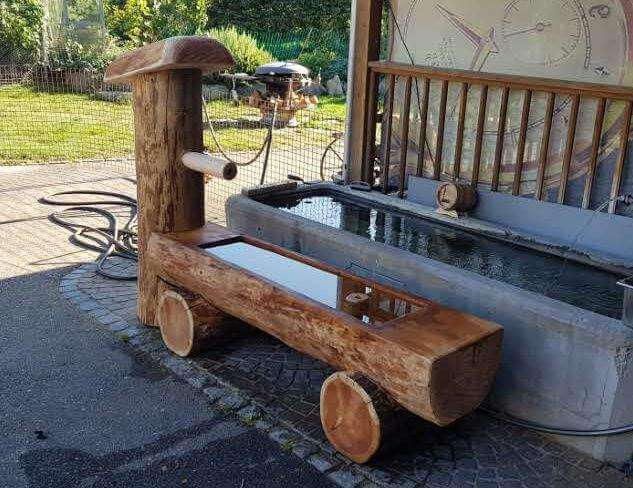 Traditioneller rustikaler Holzbrunnen im Freien mit Wasserausguss und dekorativen Elementen neben einem Wasserbecken