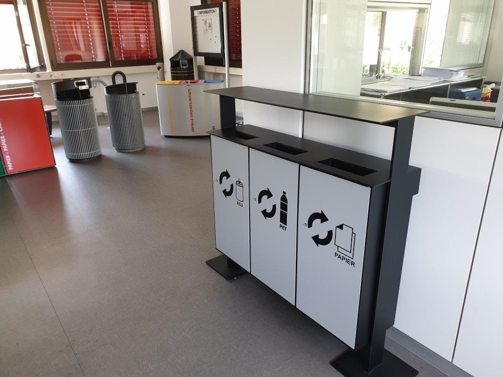 Un collecteur de déchets recyclables moderne avec trois poubelles séparées pour l'aluminium, le PET et le papier, installé dans un bureau avec des fenêtres et d'autres meubles.