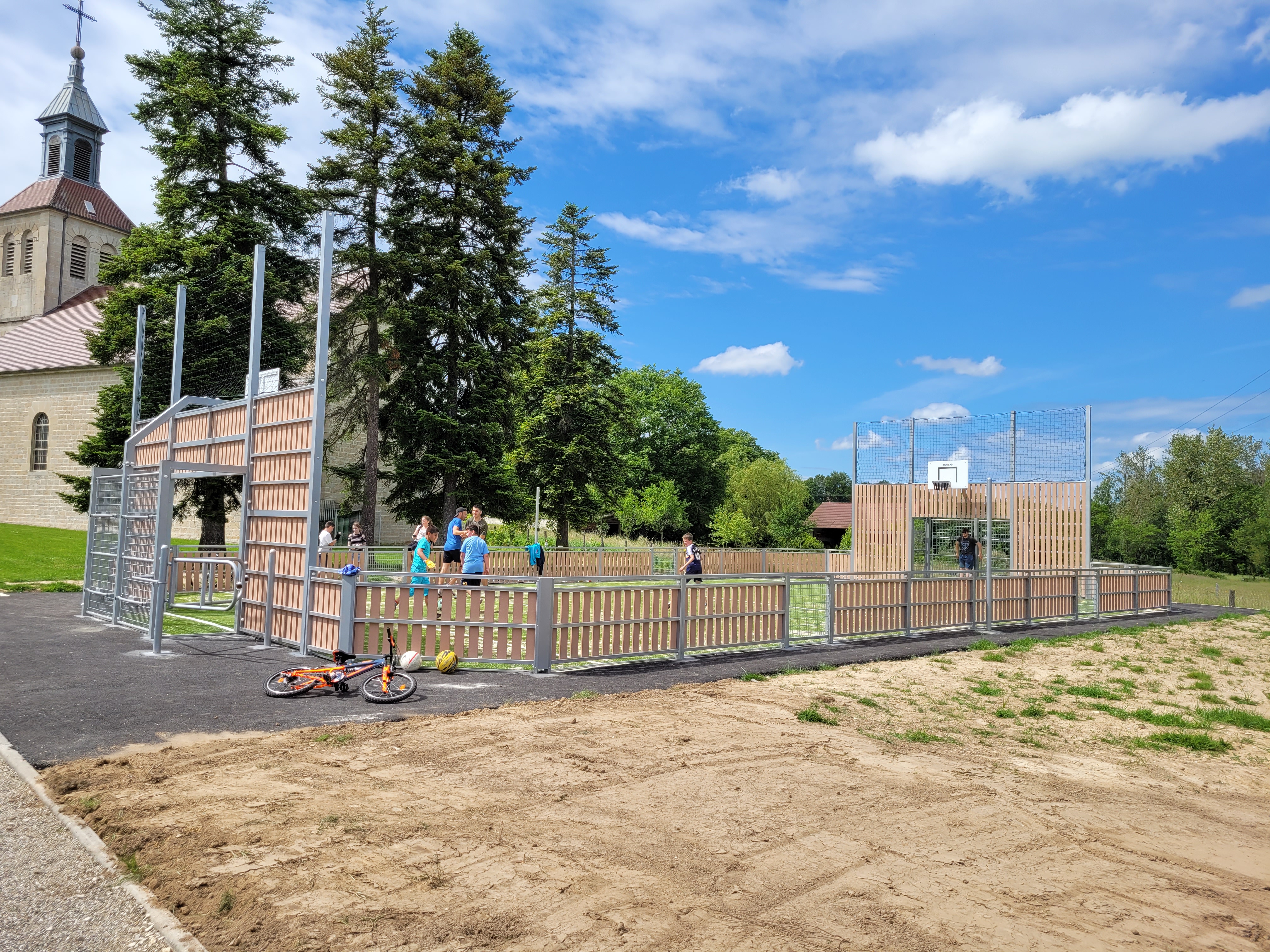 Mehrere Jugendliche spielen auf einem umzäunten Sportplatz neben einer Kirche, mit einem Fahrrad und Bällen im Vordergrund.