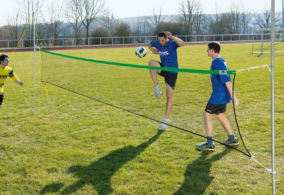Zwei Fußballspieler in blauen Trikots spielen auf einem Rasenplatz über ein Netz, während ein weiteres Teammitglied in einem gelben Trikot beobachtet.