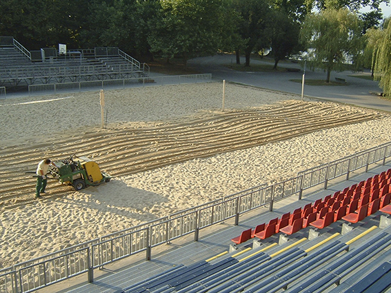 Un ouvrier entretient le sable d'un terrain de volleyball dans un stade à l'aide d'une machine ; au premier plan, des rangées de sièges vides. Des arbres en arrière-plan.