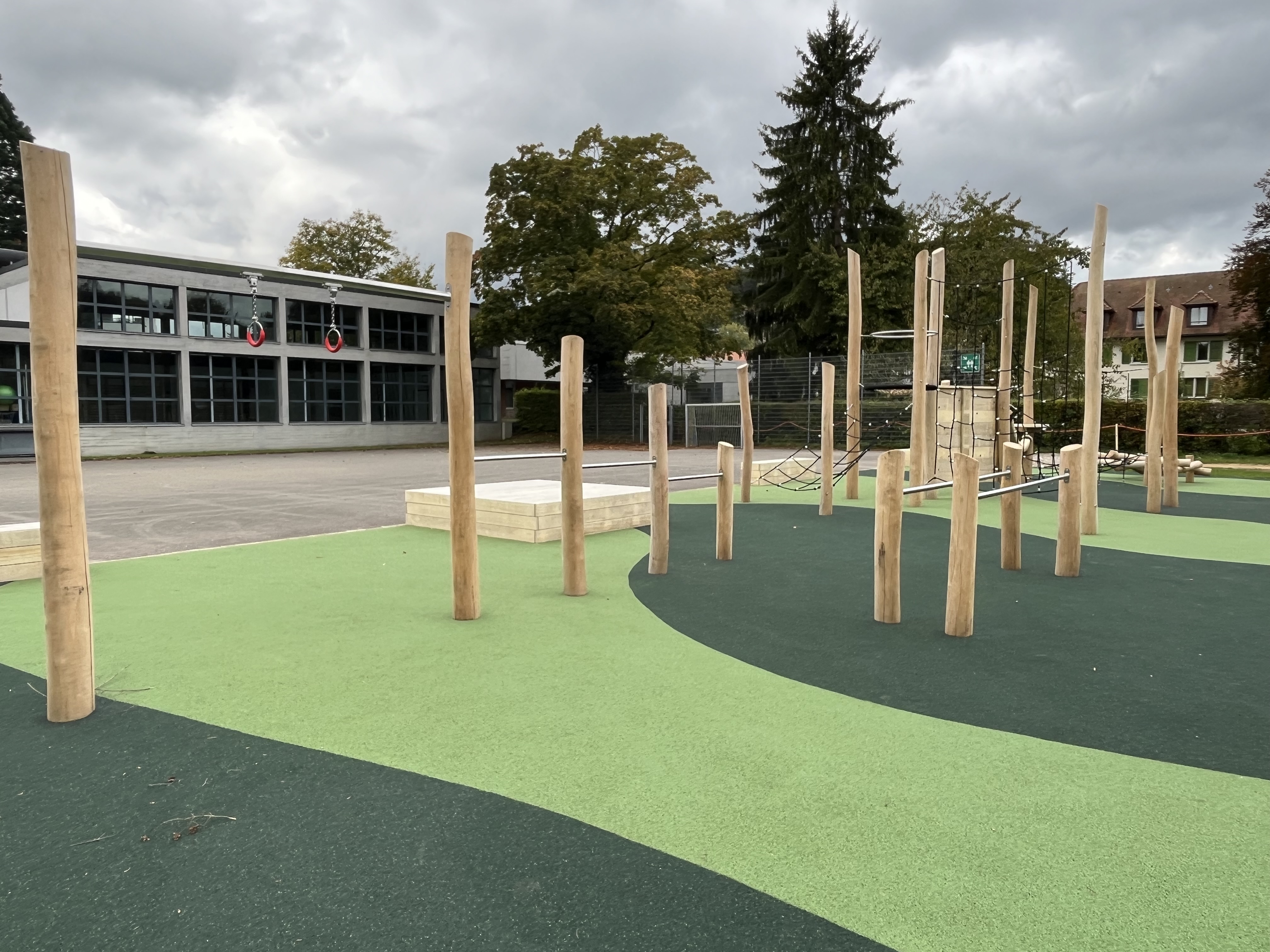 Spielplatz mit Klettergerüsten aus Holz auf grünem, wellenförmigem Bodenbelag, umgeben von Bäumen und einem Gebäude im Hintergrund