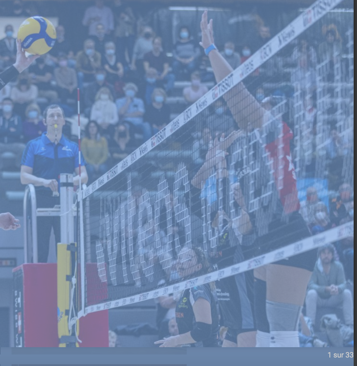 Volleyballspiel in der Halle mit Blockversuch am Netz vor Publikum.