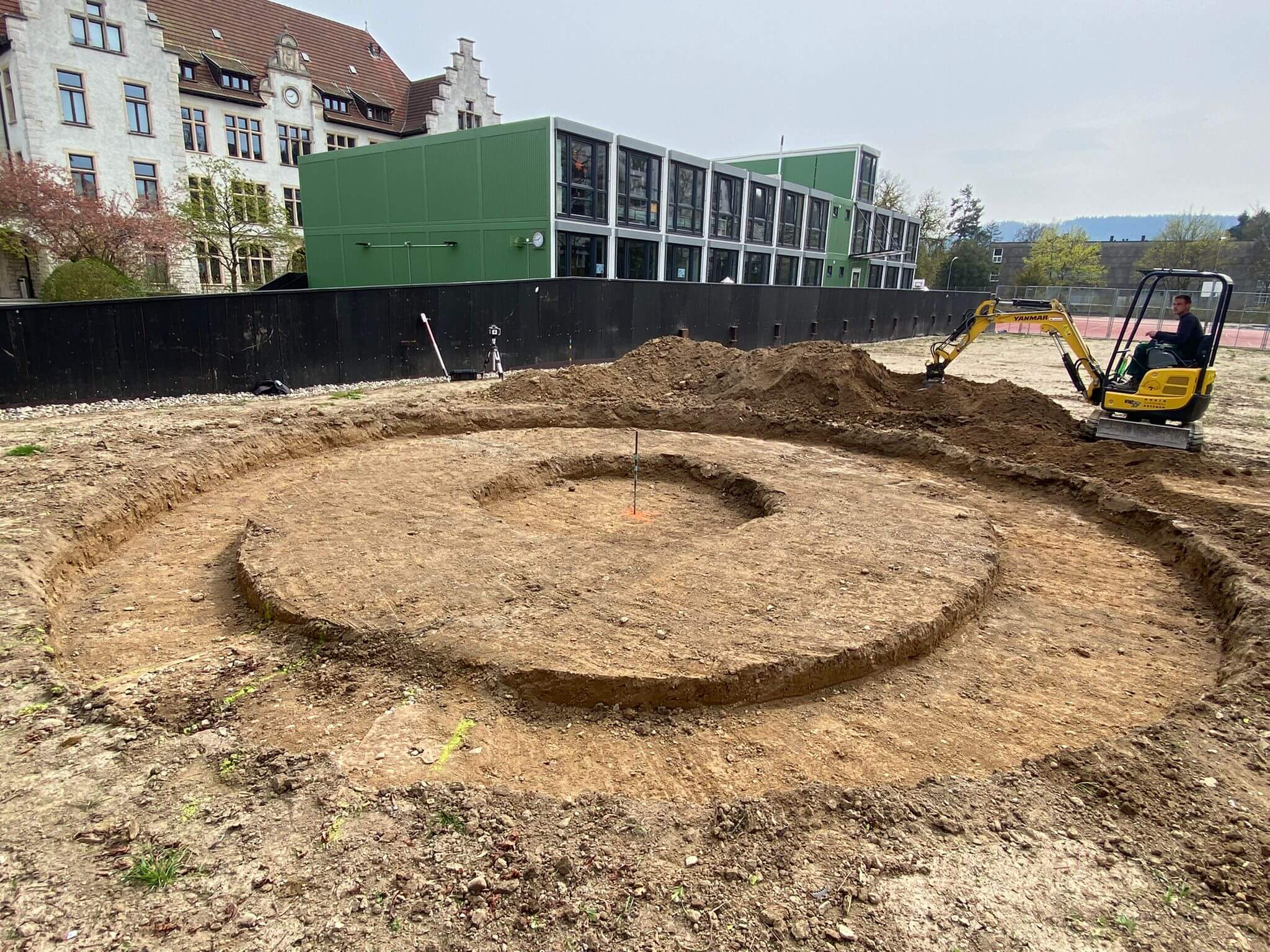 Une mini-pelle effectuant des travaux de terrassement sur un chantier, à côté d'un bâtiment moderne et d'un bâtiment historique en arrière-plan