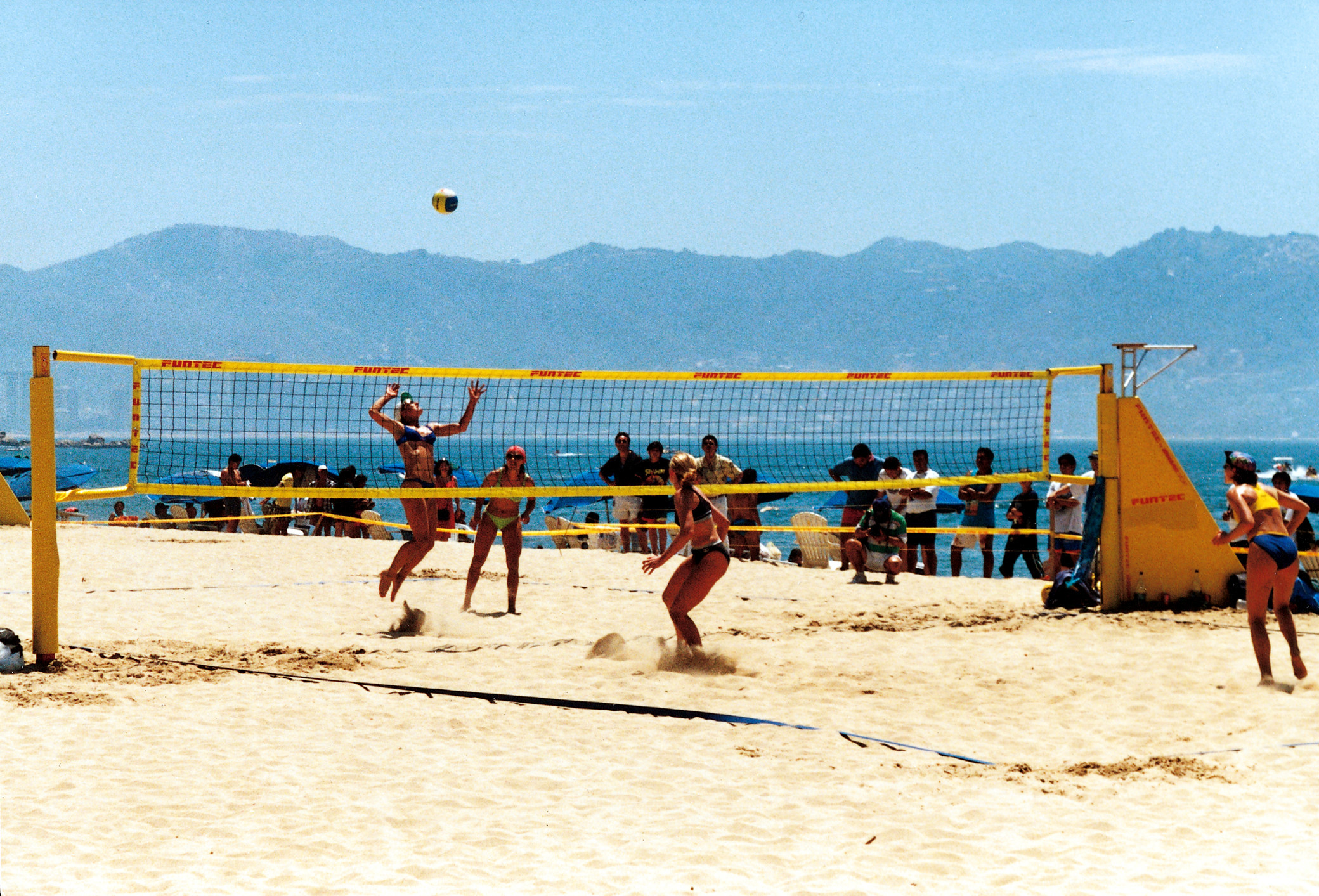 Vier Frauen beim Beachvolleyballspiel am Strand mit Zuschauern im Hintergrund, Bergen und Meer in der Ferne