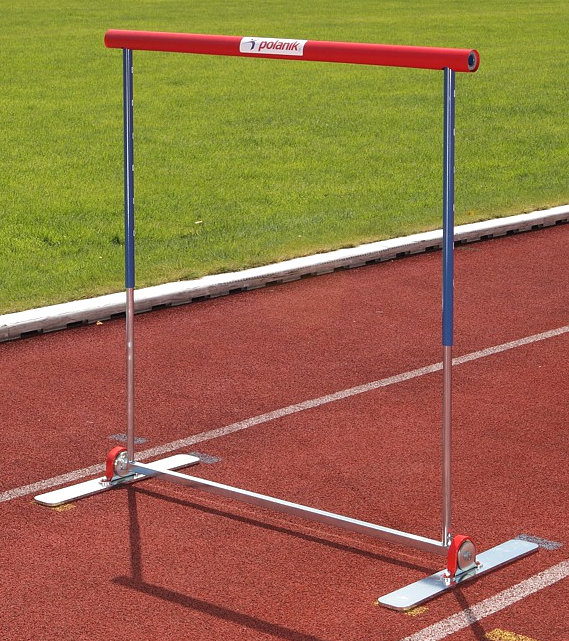 Eine rote und blaue Leichtathletik-Hürde steht auf einer Tartanbahn mit grünem Rasen im Hintergrund.