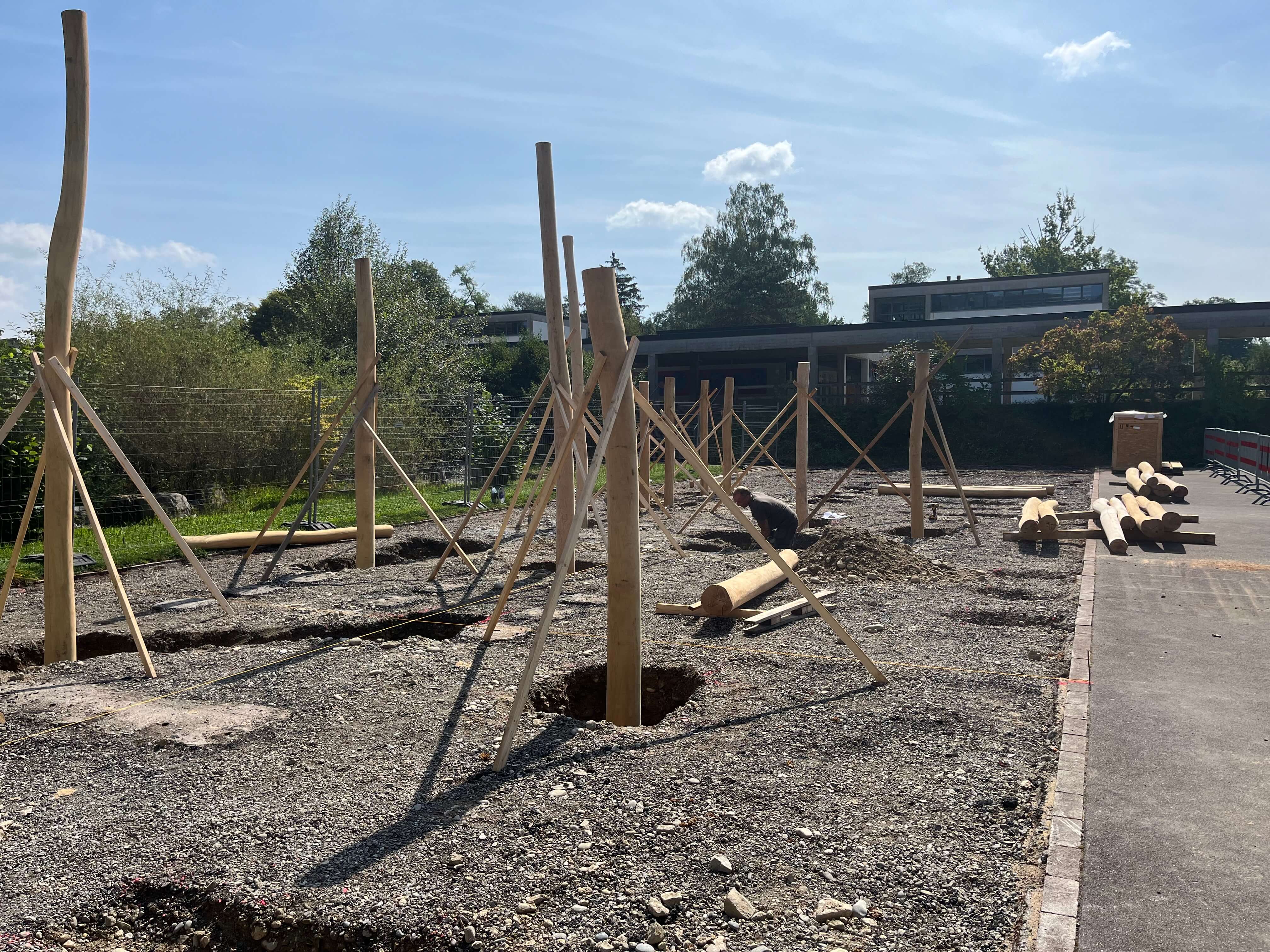 Eine Baustelle mit mehreren Holzpfählen, die auf einer Kiesfläche stehen. Im Hintergrund ist ein Gebäude und ein sonniger Himmel zu sehen.