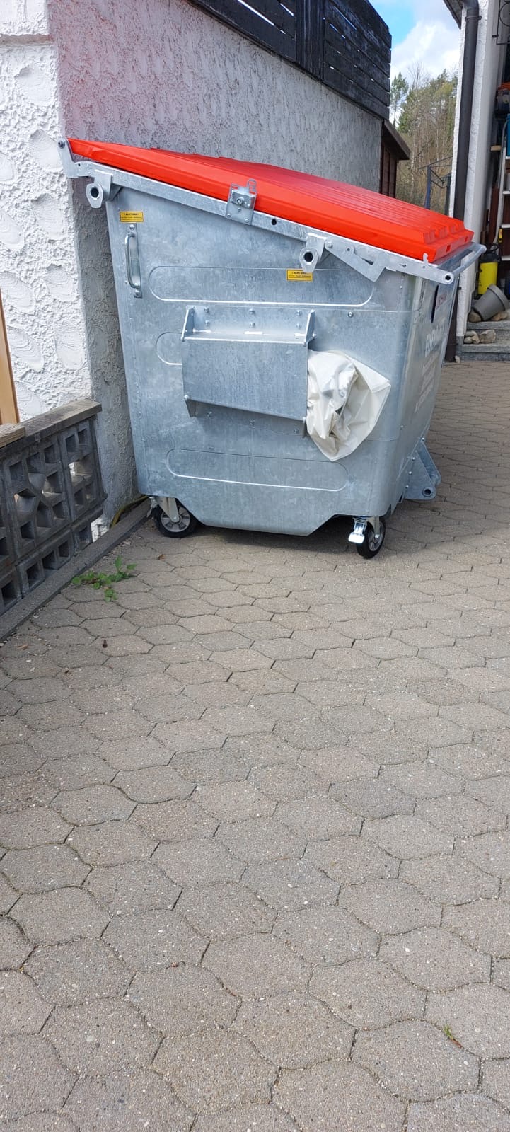 Ein großer grauer Müllcontainer mit rotem Deckel steht auf einem gepflasterten Boden und lehnt an einer weißen Wand.