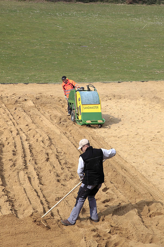 Deux personnes entretenant un terrain de jeu en sable avec une machine à nettoyer le sable et un râteau