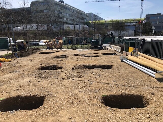 Terrain de construction avec plusieurs trous creusés et une pelleteuse en arrière-plan, entouré de bâtiments municipaux et de clôtures de chantier