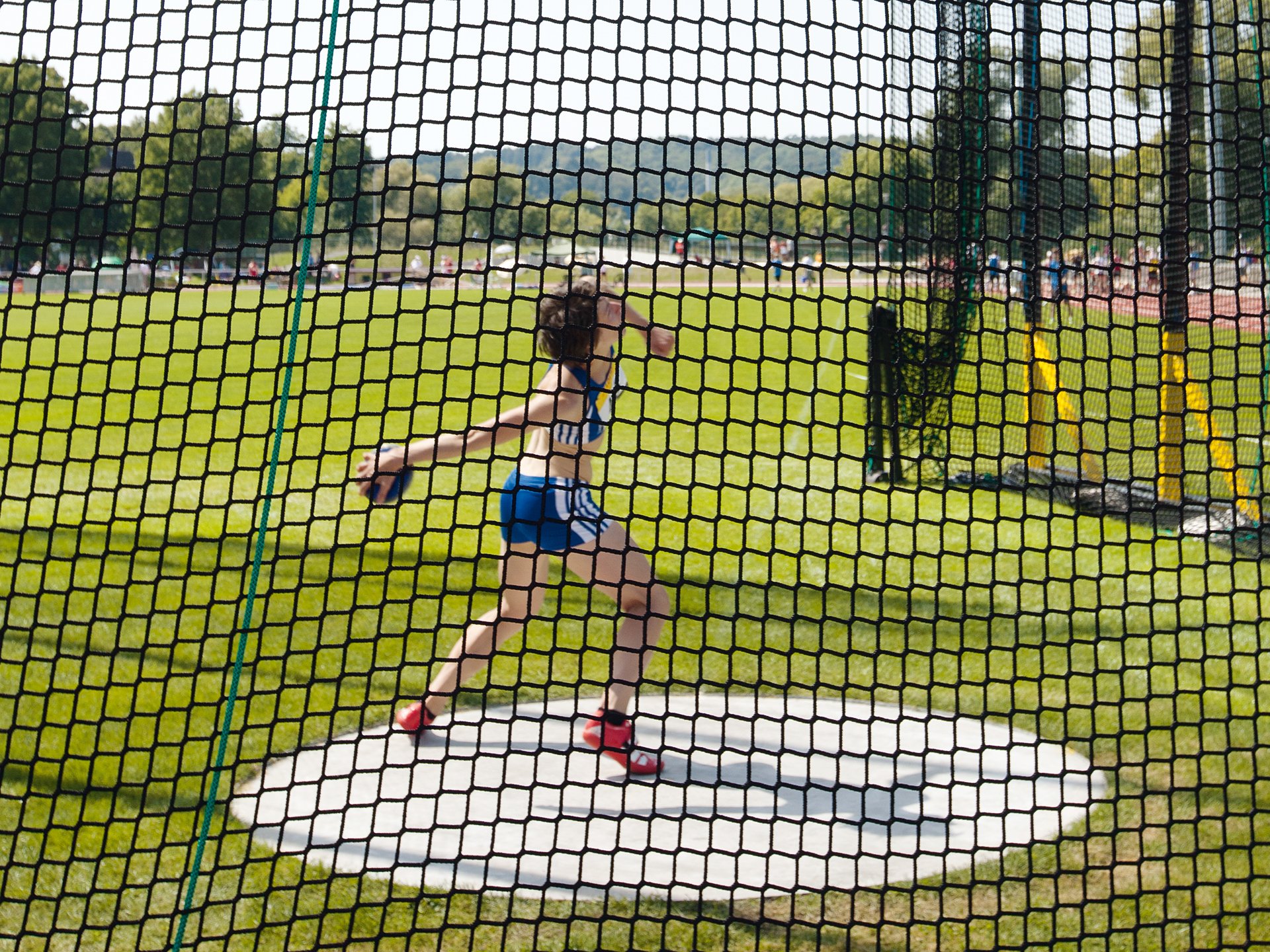 Ein Sportler in blauer Sportkleidung wirft einen Diskus innerhalb eines umzäunten Bereichs auf einem grünen Sportplatz.
