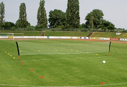 Ein mobiles Sportnetz auf einem Fußballfeld, umgeben von Markierungskegeln, mit Bäumen im Hintergrund.