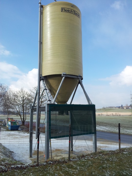 Un grand silo jaunâtre se trouve sur un terrain rural enneigé, entouré d'arbres et d'une route.