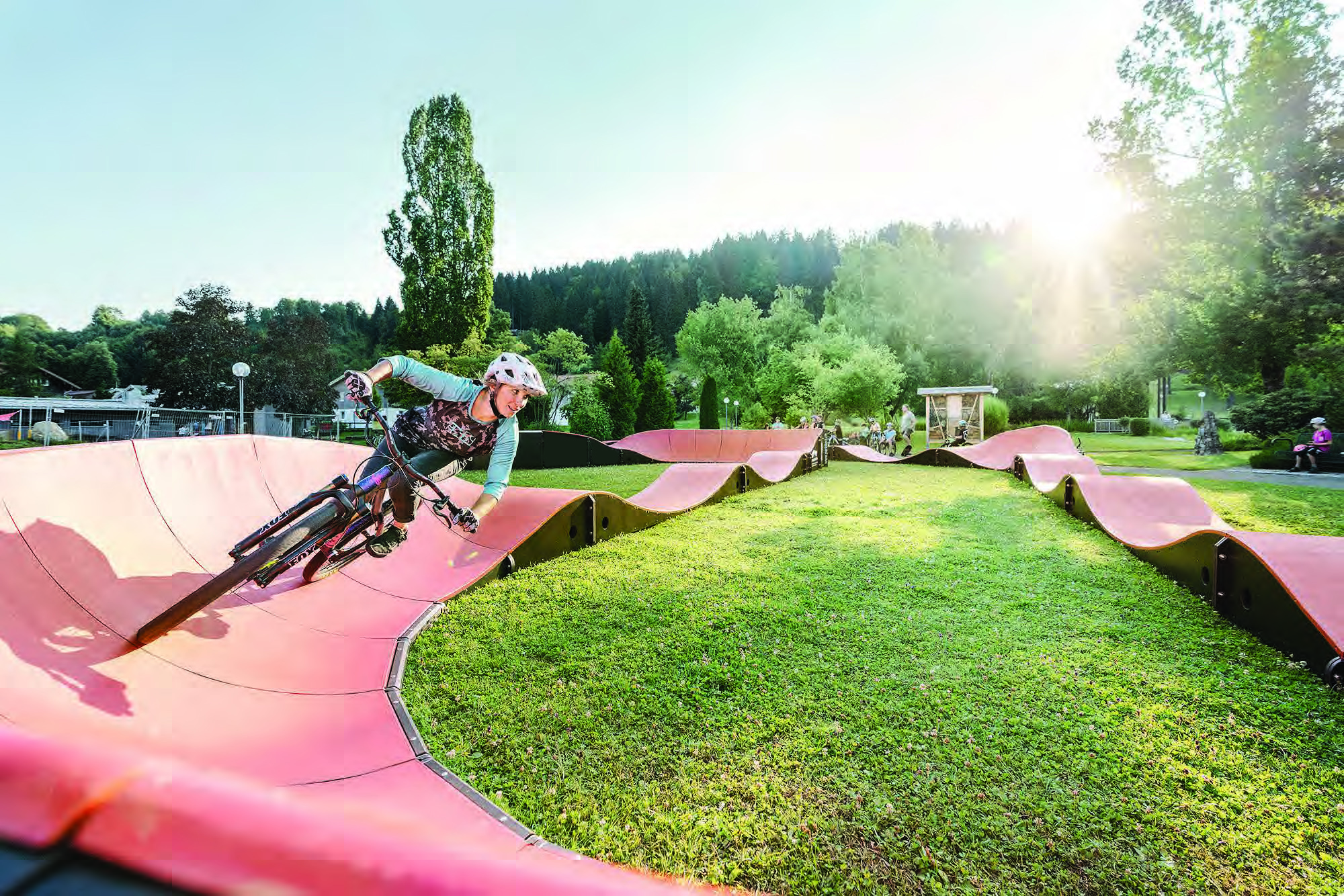 Ein Mountainbiker mit Helm auf einem Pumptrack in einer grünen Landschaft, im Hintergrund Bäume und Menschen