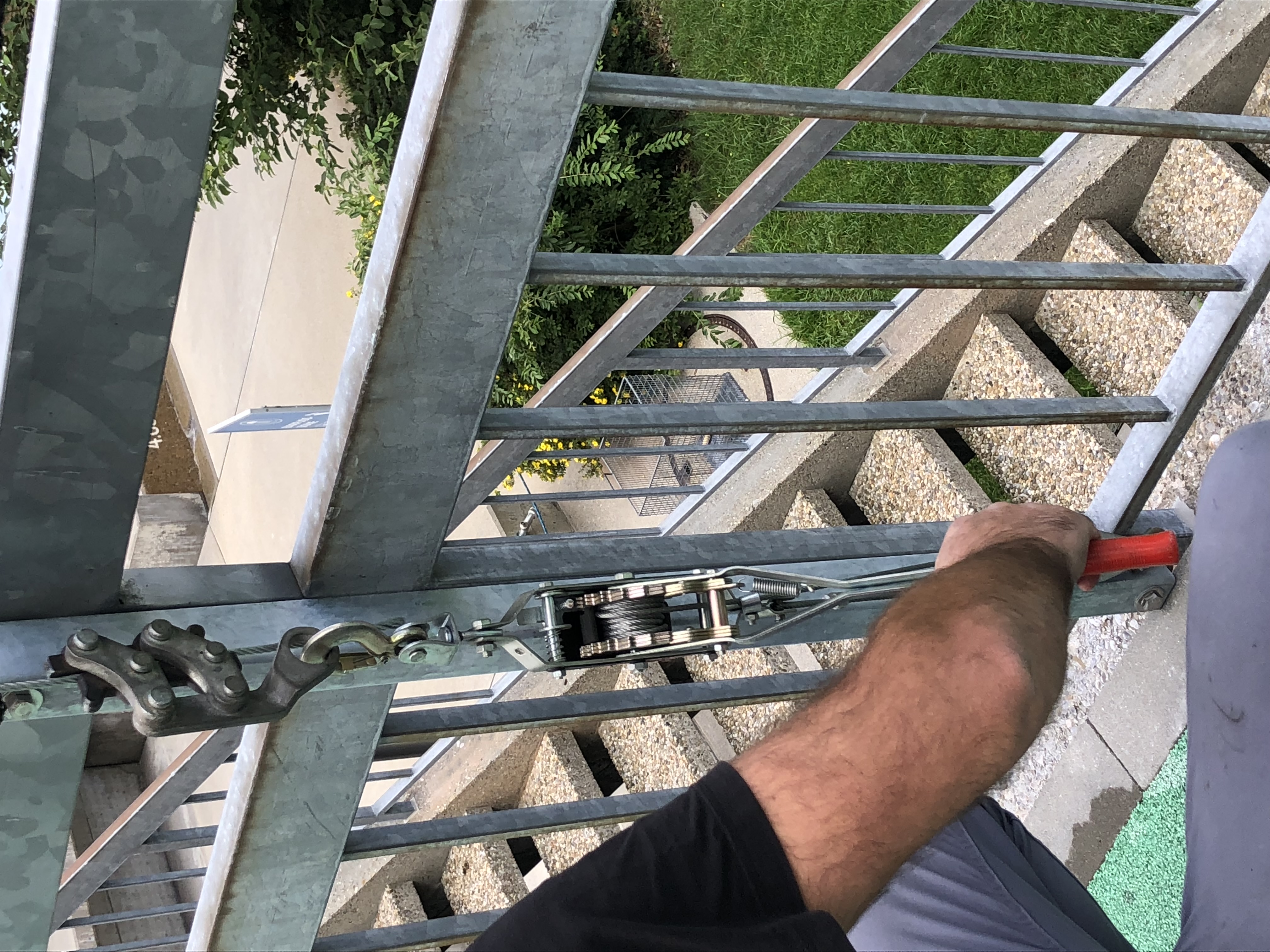 Ein Spannschloss mit Seilzug an einem Metallgeländer befestigt, im Hintergrund eine Treppe und ein Garten