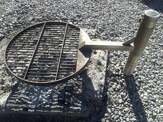 Ein runder Grillrost mit Metallstangen auf einem Untergrund aus Kieselsteinen, verbunden mit einem Metallstück.