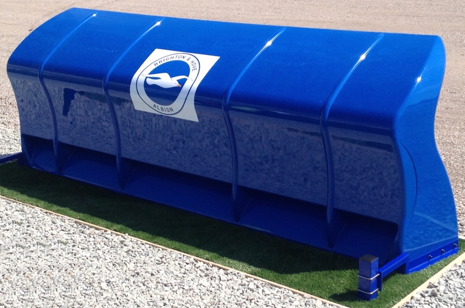 Eine blaue Kunststoffbank mit dem Logo von Brighton & Hove Albion, auf einer Fläche mit Kies und Gras platziert.