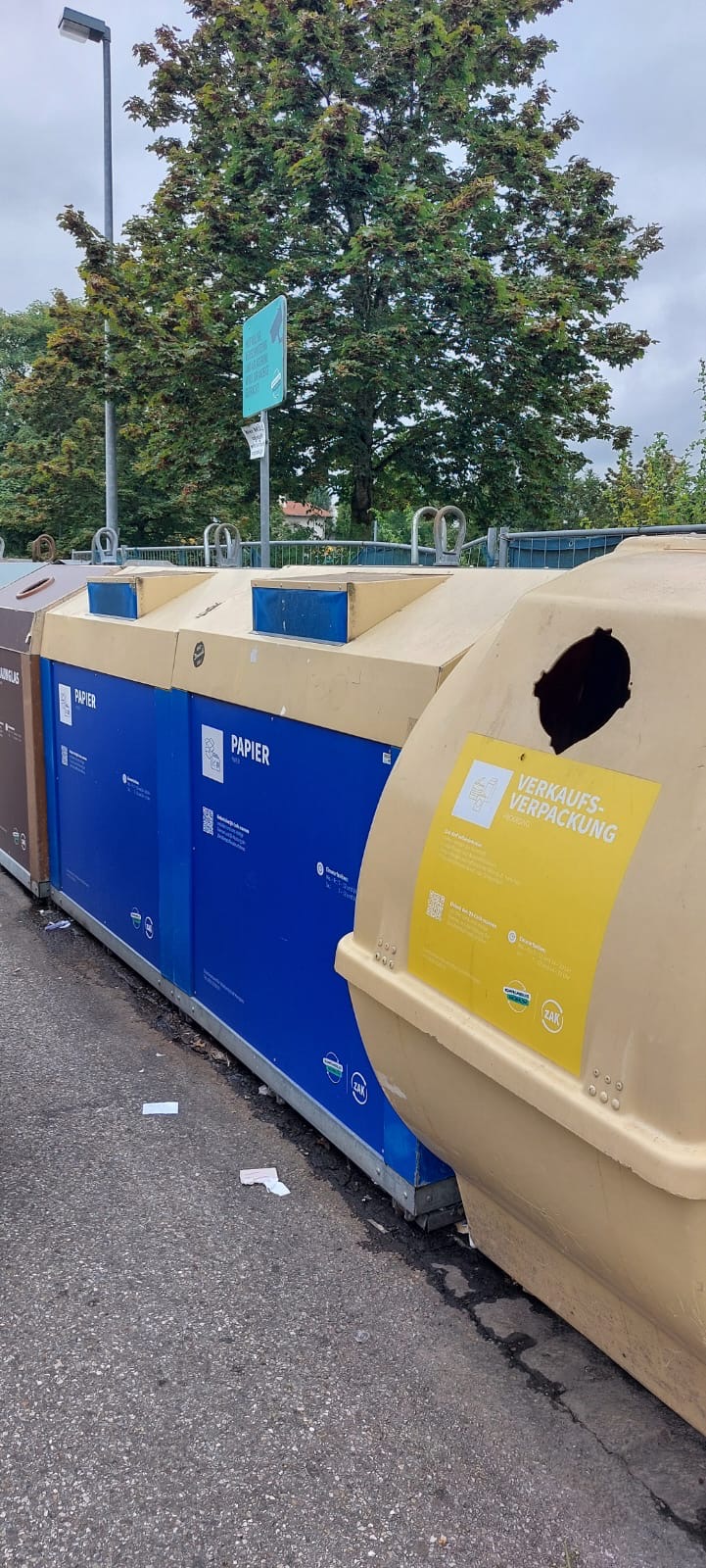 Mehrere Recyclingcontainer für Papier und Verkaufsverpackungen auf einer Straße mit Bäumen im Hintergrund.