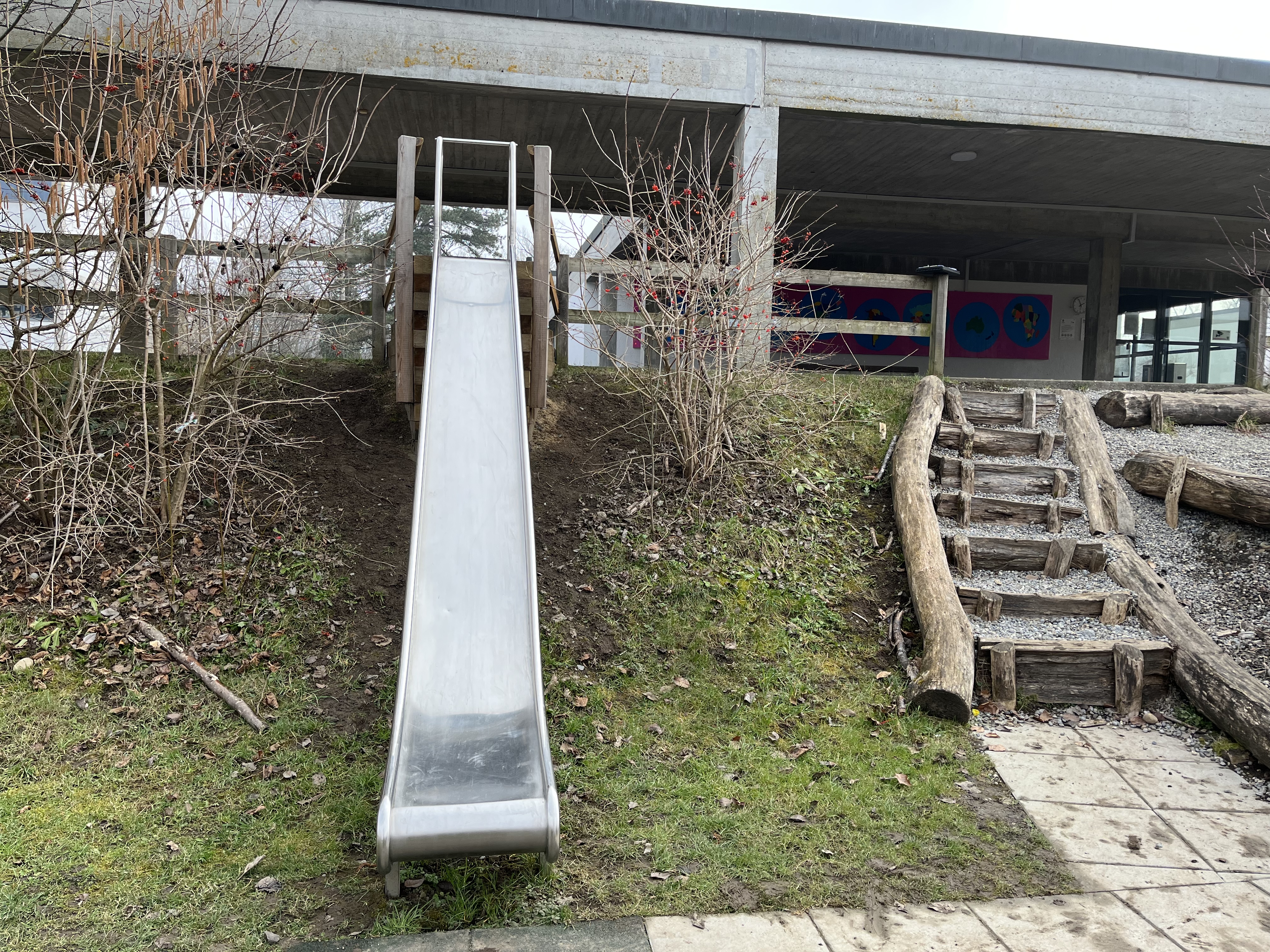 Eine Metallrutsche, die von einer kleinen Anhöhe hinunterführt, neben einer Holztreppe im Außenbereich eines Schulgebäudes.