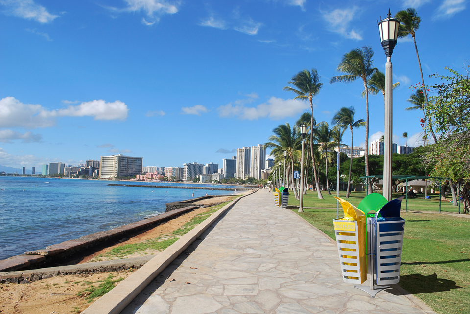 Eine Strandpromenade mit Palmen und Mülleimern entlang der Küste einer Stadt, die im Hintergrund zu sehen ist.